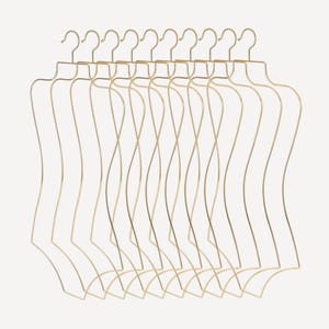 Könnte beinhalten: Eine Ansammlung goldfarbener Draht-Kleiderbügel in Form von Büsten. Die Bügel sind in einer Reihe angeordnet, jeder mit einer geschwungenen Silhouette eines Torsos und einem Haken zum Aufhängen. Der Hintergrund ist einfarbig und neutral.