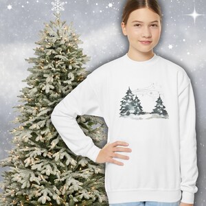 May include: A white crew neck sweatshirt with a watercolour design of two evergreen trees against a snowy backdrop. The long-sleeved top features a winter scene. A decorated Christmas tree is in the background.