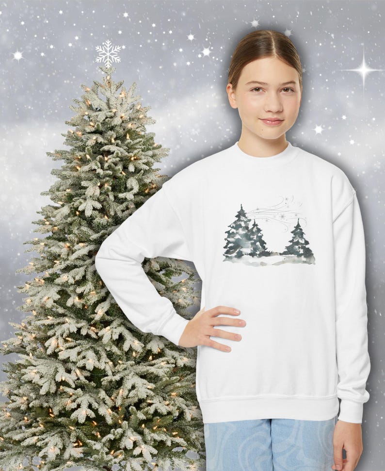 May include: A white crew neck sweatshirt with a watercolour design of two evergreen trees against a snowy backdrop. The long-sleeved top features a winter scene. A decorated Christmas tree is in the background.