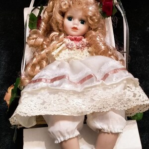 Pode incluir: Uma boneca de porcelana com cabelo loiro encaracolado, vestindo um vestido branco com detalhes e renda rosa, sentada em um balan&ccedil;o branco. A boneca tem olhos azuis e um blush rosa nas bochechas.