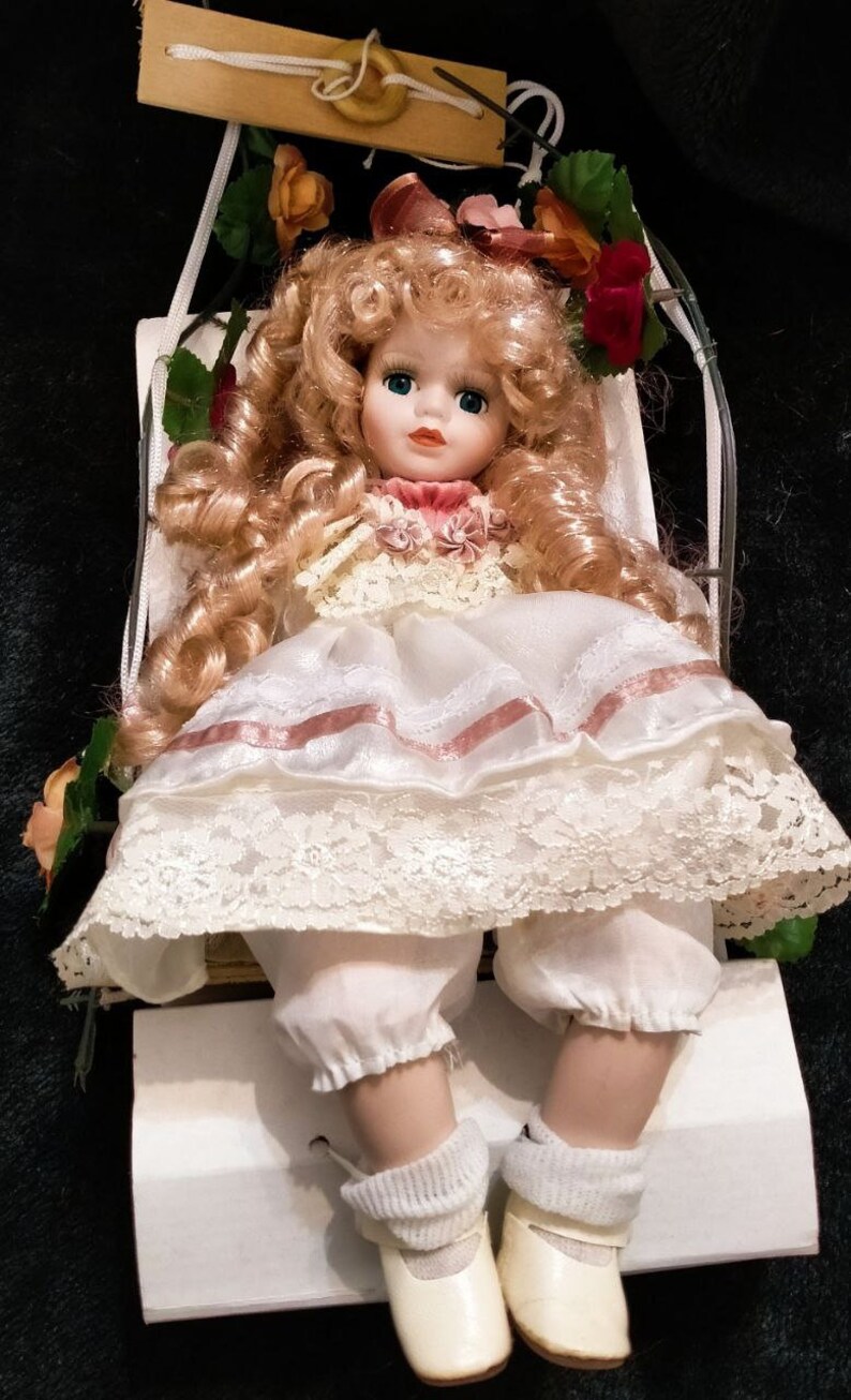 Pode incluir: Uma boneca de porcelana com cabelo loiro encaracolado, vestindo um vestido branco com detalhes e renda rosa, sentada em um balan&ccedil;o branco. A boneca tem olhos azuis e um blush rosa nas bochechas.