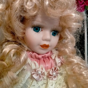 Pode incluir: Uma boneca de porcelana com cabelo loiro encaracolado vestindo um vestido de renda branco com gola rosa e detalhes de p&eacute;rolas.