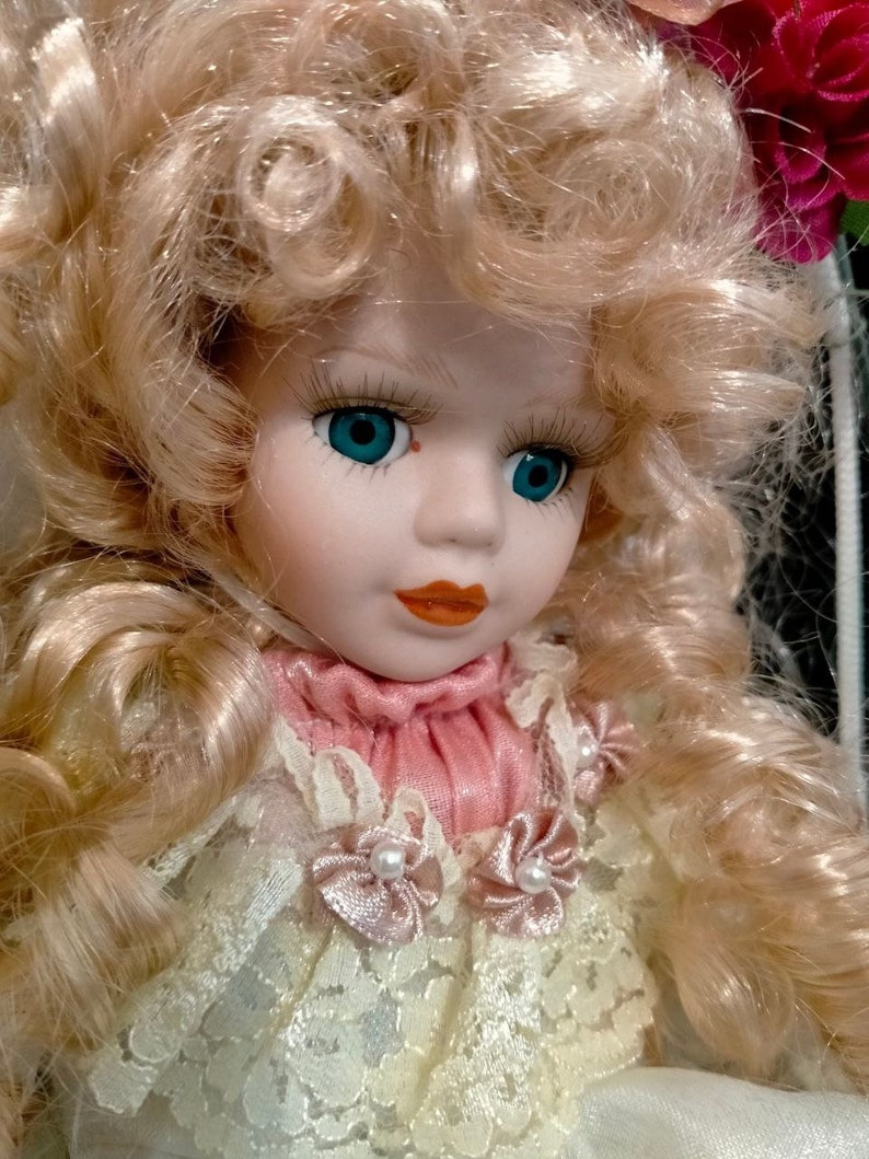 Pode incluir: Uma boneca de porcelana com cabelo loiro encaracolado vestindo um vestido de renda branco com gola rosa e detalhes de p&eacute;rolas.