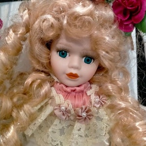 Pode incluir: Uma boneca de porcelana com cabelo loiro encaracolado, olhos azuis e um vestido rosa e branco com acabamento em renda. A boneca est&aacute; usando um la&ccedil;o rosa e est&aacute; sentada em uma cadeira com um design floral.