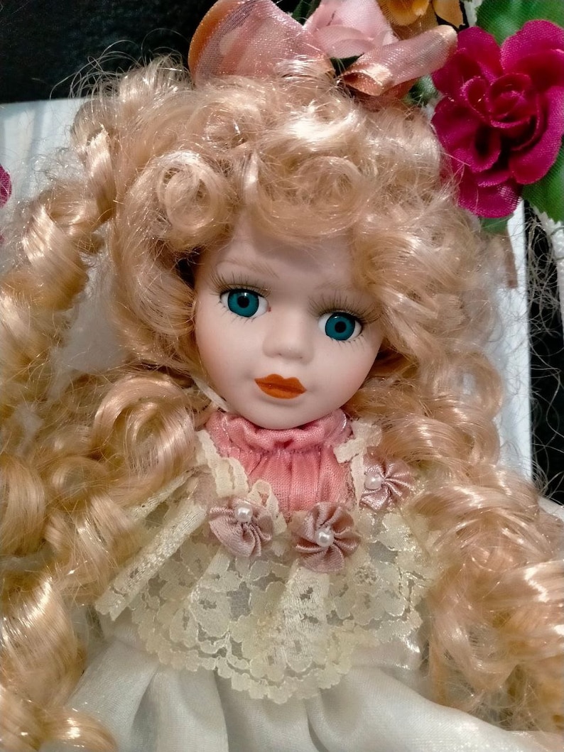 Pode incluir: Uma boneca de porcelana com cabelo loiro encaracolado, olhos azuis e um vestido rosa e branco com acabamento em renda. A boneca est&aacute; usando um la&ccedil;o rosa e est&aacute; sentada em uma cadeira com um design floral.