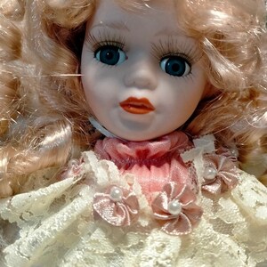 Pode incluir: Uma boneca de porcelana com cabelo loiro encaracolado, vestindo um vestido de renda branca com detalhes em cetim rosa e enfeites de p&eacute;rolas. A boneca tem olhos azuis e um sorriso pintado.