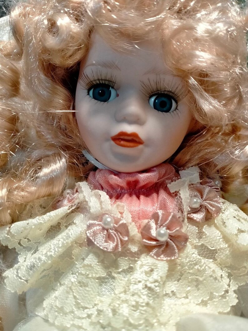 Pode incluir: Uma boneca de porcelana com cabelo loiro encaracolado, vestindo um vestido de renda branca com detalhes em cetim rosa e enfeites de p&eacute;rolas. A boneca tem olhos azuis e um sorriso pintado.