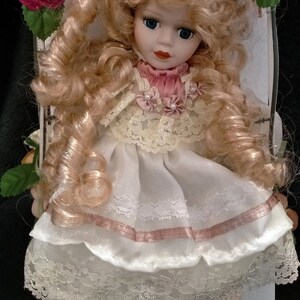 Pode incluir: Uma boneca de porcelana com cabelo loiro encaracolado, vestindo um vestido branco com detalhes rosa e acabamento em renda. A boneca est&aacute; sentada em uma superf&iacute;cie branca com rosas de cada lado.