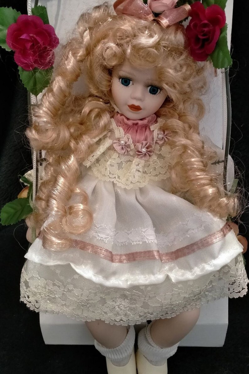 Pode incluir: Uma boneca de porcelana com cabelo loiro encaracolado, vestindo um vestido branco com detalhes rosa e acabamento em renda. A boneca est&aacute; sentada em uma superf&iacute;cie branca com rosas de cada lado.