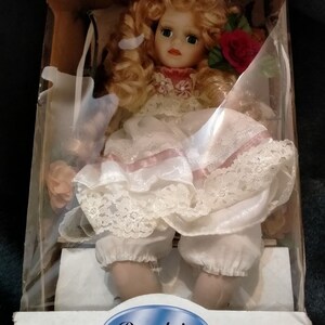 Pode incluir: Uma boneca de porcelana com cabelo loiro e um vestido branco com detalhes rosa. A boneca est&aacute; sentada em uma cadeira e segurando uma rosa vermelha. A caixa diz "Porcelain Doll".