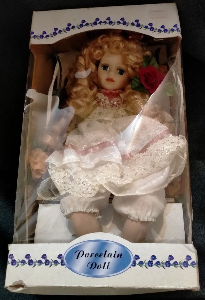 Pode incluir: Uma boneca de porcelana com cabelo loiro e um vestido branco com detalhes rosa. A boneca est&aacute; sentada em uma cadeira e segurando uma rosa vermelha. A caixa diz "Porcelain Doll".