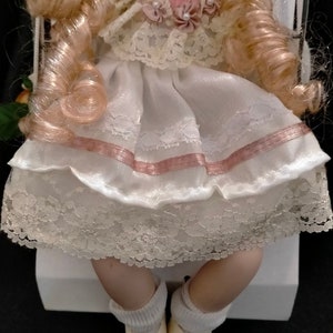Pode incluir: Uma boneca de porcelana vestindo um vestido branco com detalhes em rosa e renda. A boneca tem cabelo loiro encaracolado e est&aacute; usando meias e sapatos brancos.