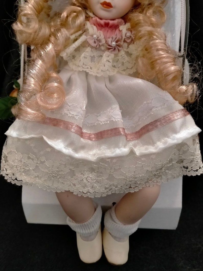 Pode incluir: Uma boneca de porcelana vestindo um vestido branco com detalhes em rosa e renda. A boneca tem cabelo loiro encaracolado e est&aacute; usando meias e sapatos brancos.