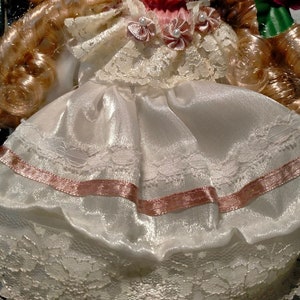 Pode incluir: Uma boneca de porcelana com cabelo loiro vestindo um vestido branco com detalhes de fita rosa e acabamento em renda. A boneca est&aacute; segurando uma rosa vermelha.