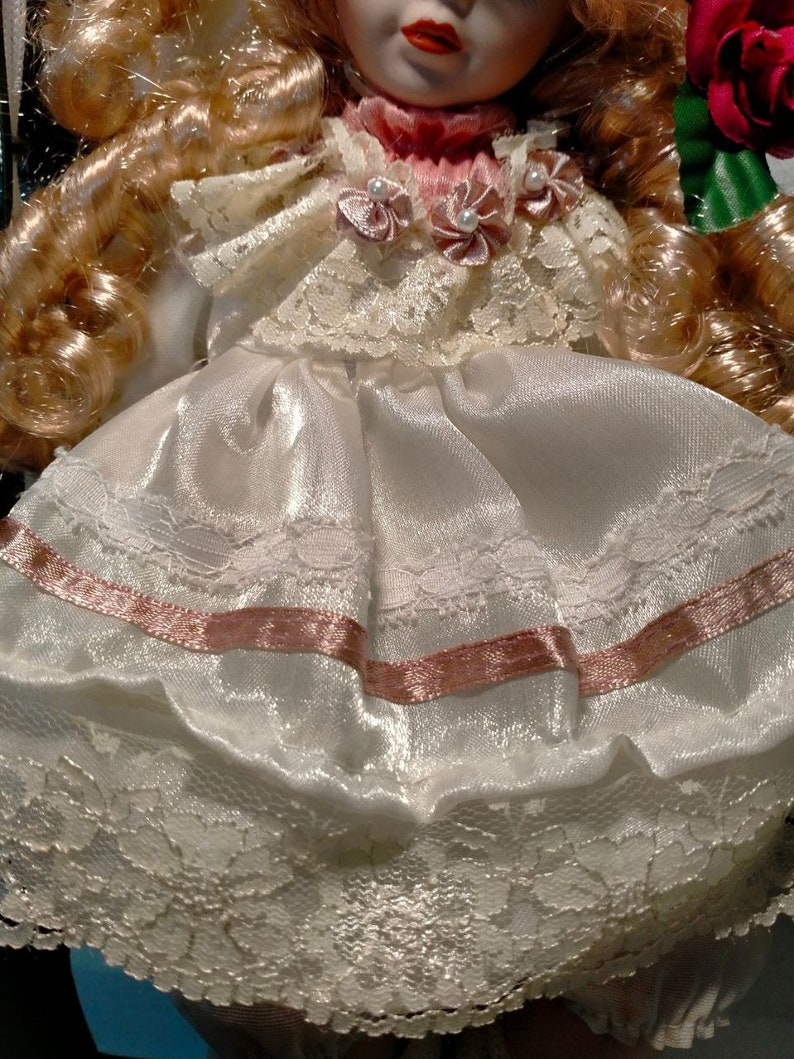 Pode incluir: Uma boneca de porcelana com cabelo loiro vestindo um vestido branco com detalhes de fita rosa e acabamento em renda. A boneca est&aacute; segurando uma rosa vermelha.