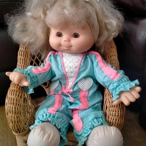 Könnte beinhalten: Eine Vintage-Puppe mit blonden Haaren und blauen Augen, bekleidet mit einem blau-rosa Outfit mit weißen Herzakzenten. Die Puppe sitzt in einem kleinen Korbstuhl, mit weißen Stofffüßen und einem weißen Band im Haar.