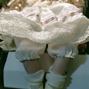 Pode incluir: Uma boneca de porcelana vestindo um vestido branco com detalhes em renda e fitas rosa. A boneca tem cabelo loiro e est&aacute; usando sapatos brancos.