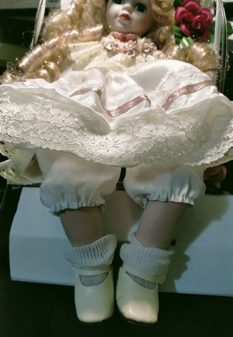 Pode incluir: Uma boneca de porcelana vestindo um vestido branco com detalhes em renda e fitas rosa. A boneca tem cabelo loiro e est&aacute; usando sapatos brancos.