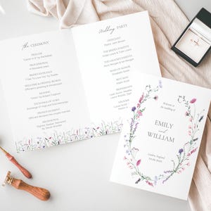 Pode incluir: Conjunto de papelaria de casamento com programas de casamento, uma caixa de anel e um selo de cera. Os programas são brancos com detalhes florais e texto. A caixa do anel é preta. Anéis de casamento dourados também são visíveis.