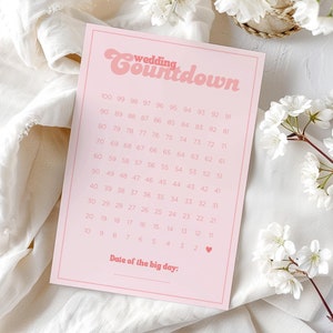 Puede incluir: Una tarjeta rosa de cuenta regresiva para bodas con la frase "wedding countdown" en una fuente retro. La tarjeta tiene una cuadrícula de números del 1 al 100, con espacio para la fecha de la boda. Flores blancas y una tela blanca en el fondo.