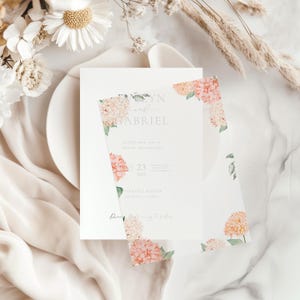 Pode incluir: Um conjunto de convites de casamento com um cartão branco com os nomes "Evelyn e Gabriel" em escrita cinza. O cartão é adornado com flores de hortênsia pêssego e rosa em aquarela. A data "23 de agosto de 2025" também é visível.