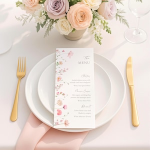 May include: A table setting with a floral arrangement, gold cutlery, and a menu card. The menu card features floral designs and the text "The MENU". The plates are white, and the napkin is pink.