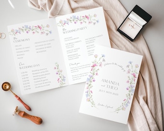 May include: A wedding invitation suite with a floral design. The invitation features a wreath of watercolor flowers in shades of pink, blue, and green. The text "Amanda and Theo" is printed in a simple font, along with the date "July 19th, 2023" and the location "London, England". The matching program includes a list of the wedding party and the ceremony and reception schedule.