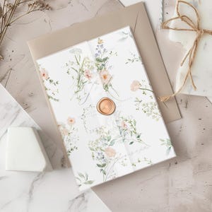 Può includere: Un invito di nozze con un design floreale ad acquerello, con fiori color pesca e lavanda, foglie verdi e un sigillo di cera dorata. L'invito è su sfondo bianco con testo. C'è una busta beige dietro l'invito.