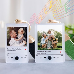 May include: Two white photo frames with a music player design. Each frame features a photo of a family. The frames are attached to keychains with green and black leather straps. The text on the frames reads "Dance With My Father" and "Luther Vandross".