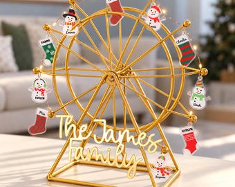 May include: A gold-colored metal ferris wheel display stand featuring the text The James Family in script font. The wheel is adorned with small LED string lights and various hanging acrylic ornaments shaped like snowmen and Christmas stockings.