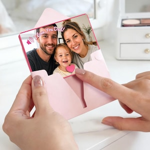 May include: A light pink envelope-shaped card featuring a photograph of a man, a woman, and a young child. The card includes the text "I Love You Dad!" with a speaker icon. The design incorporates a small pink heart accent on the child's shirt.