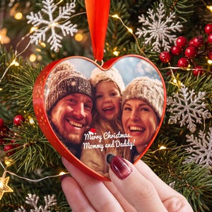 Op de afbeelding: Een hartvormige rode kerstversiering met een afgedrukte foto van een lachend gezin met wintermutsen op. Het ornament bevat de witte tekst Merry Christmas, Mommy and Daddy! met een klein rood kerstmutssymbool boven de belettering.