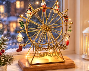 May include: A gold-colored metal Ferris wheel ornament display stand on a rectangular wooden base. The structure features string lights and hanging acrylic charms shaped like snowmen and Christmas stockings, with the text The James Family.