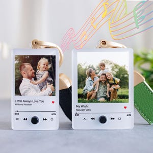 May include: Two rectangular acrylic keychains featuring custom photo prints styled like music player interfaces. The left keychain displays a photo of a man and child with text I Will Always Love You by Whitney Houston. The right shows a family photo with text My Wish by Rascal Flatts, both with gold hardware.