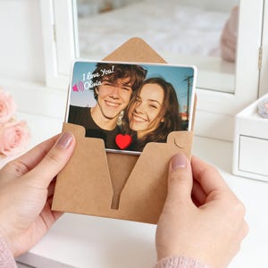 Könnte beinhalten: Eine rechteckige Karte mit einem Foto eines lächelnden Paares wird in einem braunen Papierumschlag gehalten. Die Karte trägt den Text "I love you! Olivia" und ein rotes Herz. Der Umschlag befindet sich vor einem weißen Hintergrund.