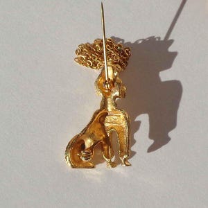 Puede incluir: Un broche de metal dorado con forma de caniche sentado y acabado texturizado. La pieza cuenta con un mecanismo de alfiler largo en la parte posterior y un elemento de cadena decorativa dorada unido a la parte superior de la cabeza del caniche.