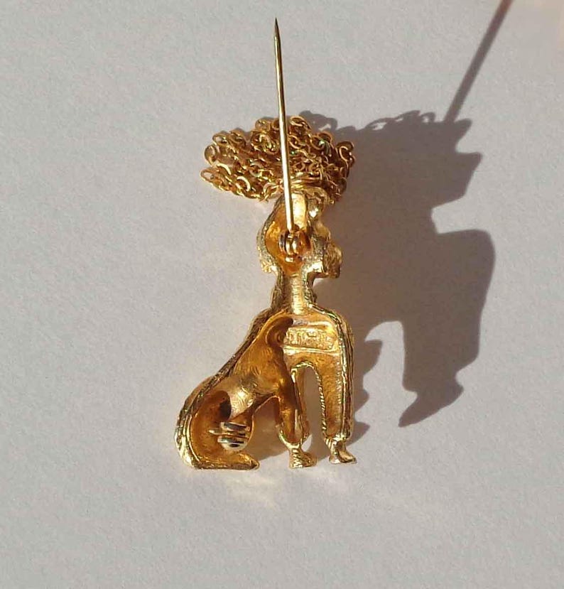 Puede incluir: Un broche de metal dorado con forma de caniche sentado y acabado texturizado. La pieza cuenta con un mecanismo de alfiler largo en la parte posterior y un elemento de cadena decorativa dorada unido a la parte superior de la cabeza del caniche.