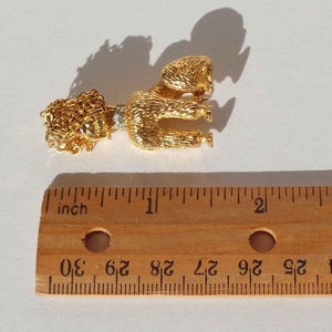 Puede incluir: Un broche de caniche en tono dorado con acabado texturizado, que presenta un peque&ntilde;o collar con piedras transparentes. El caniche est&aacute; colocado sobre una regla de madera, mostrando que el broche mide aproximadamente 4,5 cm de largo total.
