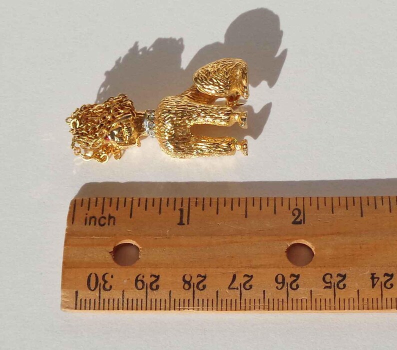Puede incluir: Un broche de caniche en tono dorado con acabado texturizado, que presenta un peque&ntilde;o collar con piedras transparentes. El caniche est&aacute; colocado sobre una regla de madera, mostrando que el broche mide aproximadamente 4,5 cm de largo total.