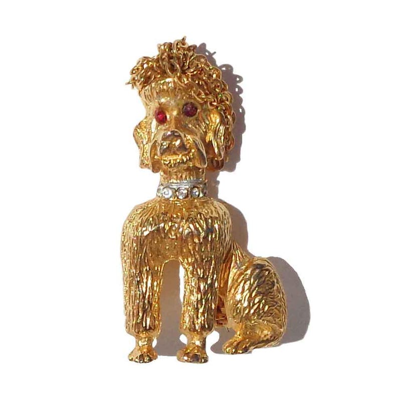 Puede incluir: Un broche de caniche en tono dorado con pelaje texturizado, dos peque&ntilde;os ojos de piedra roja y un collar en tono plateado adornado con una hilera de brillantes piedras de estr&aacute;s transparentes. El perro aparece sentado.