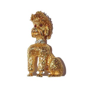 Puede incluir: Un broche de caniche en tono dorado con pelaje texturizado, un collar con pedrer&iacute;a transparente y dos peque&ntilde;as piedras rojas para los ojos. El perro est&aacute; representado en posici&oacute;n sentada con la cabeza ligeramente girada hacia un lado.
