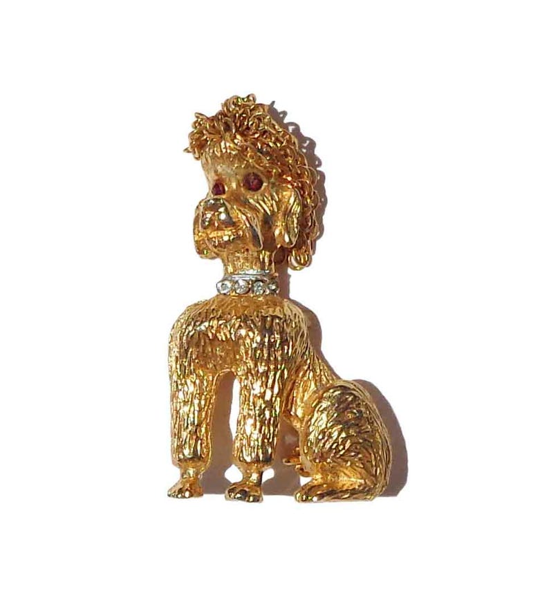 Puede incluir: Un broche de caniche en tono dorado con pelaje texturizado, un collar con pedrer&iacute;a transparente y dos peque&ntilde;as piedras rojas para los ojos. El perro est&aacute; representado en posici&oacute;n sentada con la cabeza ligeramente girada hacia un lado.