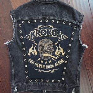 May include: Black denim vest with a distressed look. The back features a black panel with the gold text "KROKUS" above a sugar skull design and the phrase "YOU NEVER ROCK ALONE". Metal grommets line the edges.