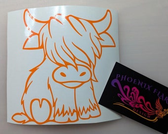 May include: Orange vinyl decal of a cartoon Highland cow with a heart-shaped patch on its side. The decal is on a white background. The text "PHOENIX FLARE DESIGN" is on a black background.