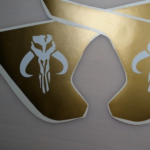 May include: Two gold vinyl decals with a white Mandalorian symbol. The symbol is a skull with horns and a vertical line down the center.
