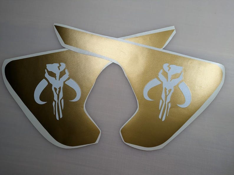 May include: Two gold vinyl decals with a white Mandalorian symbol. The symbol is a skull with horns and a vertical line down the center.