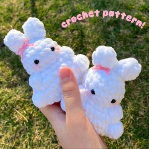 Op de afbeelding: Twee witte gehaakte konijnen knuffels met roze strikjes. De konijnen zijn pluche en hebben zwarte ogen. De tekst "crochet pattern!" is geschreven in roze letters op een groene achtergrond.