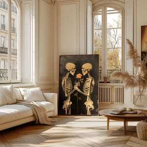 May include: A framed artwork featuring two golden-hued skeletons holding a rose against a dark backdrop. The skeletons' rib cages and skulls are detailed. The artwork is displayed in a bright room with a white sofa and large windows, creating a contrast.