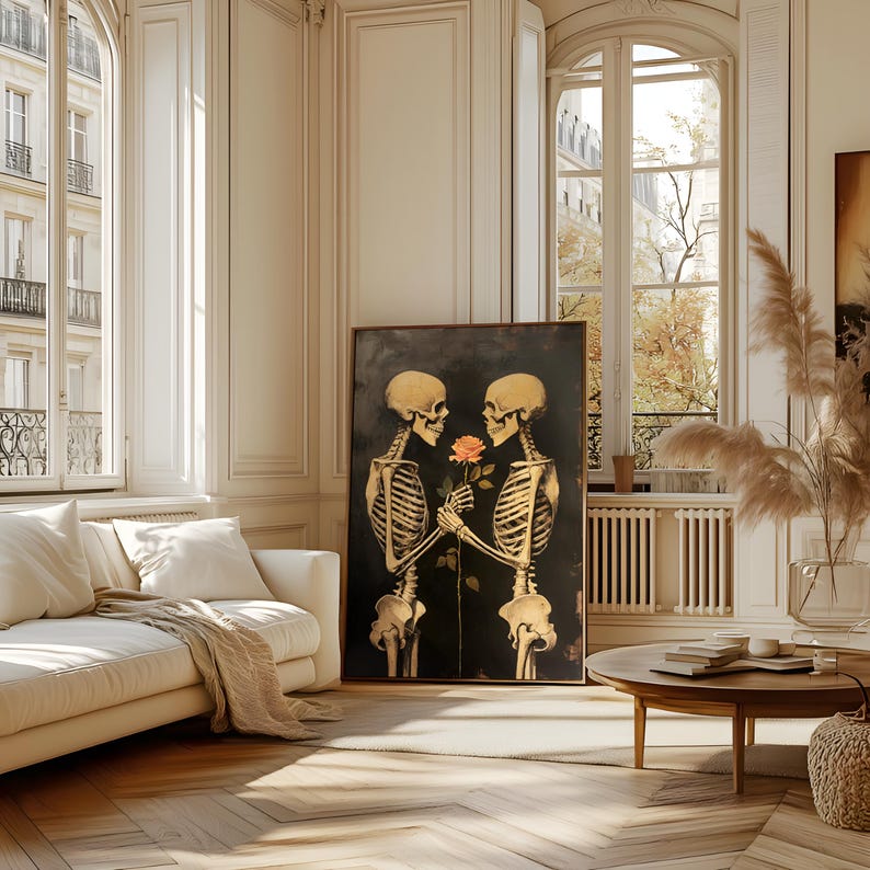 May include: A framed artwork featuring two golden-hued skeletons holding a rose against a dark backdrop. The skeletons' rib cages and skulls are detailed. The artwork is displayed in a bright room with a white sofa and large windows, creating a contrast.