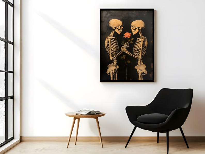May include: A framed print depicting two skeletons, rendered in sepia tones, holding a rose against a dark backdrop. The artwork is mounted on a white wall, with a black chair and a wooden side table visible in the foreground. The overall aesthetic is gothic.